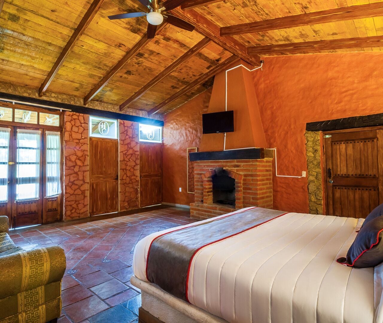 Un dormitorio con una cama grande, chimenea, paredes de piedra, puertas de madera y un ventilador de techo.