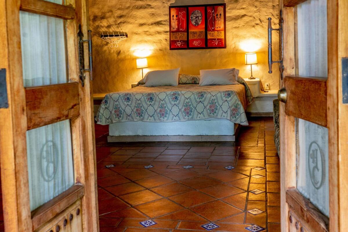 A rustic bedroom with a stone wall, a bed with a patterned bedspread, two bedside lamps, and a wooden ceiling.