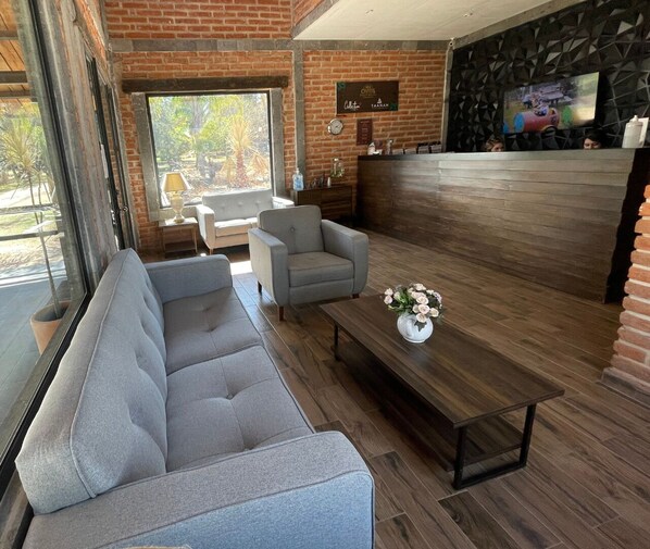 A modern lounge area with a grey sofa, a small armchair, and a wooden coffee table with a vase of flowers. - Taanah Hotel & Glamping (Zapotlanejo)