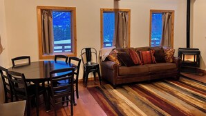 A cozy living room with a wooden floor, a brown sofa with patterned cushions, a dining table with chairs, and a wood-burning stove. - Cozy Woodland Escape: Scenic Cabin Retreat in Alpine, WY (Alpine)