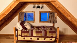 A loft bedroom with a wooden ceiling, two windows with curtains, a bed with a patterned quilt, and a small table with two glasses. - Cozy Woodland Escape: Scenic Cabin Retreat in Alpine, WY (Alpine)