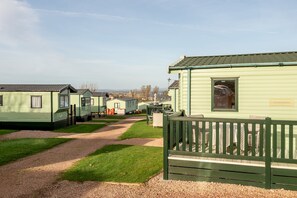 A row of green mobile homes with a gravel path leading to a fenced area with tables and chairs.