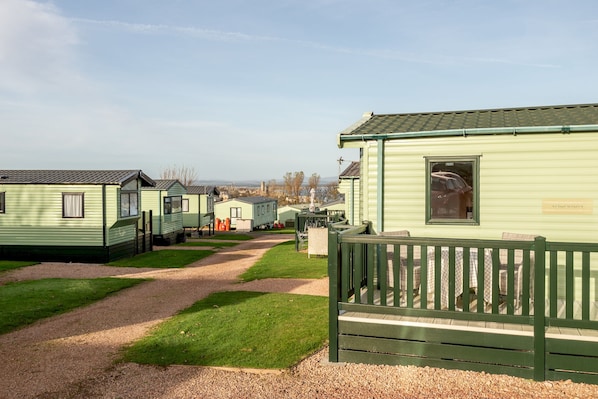 A row of green mobile homes with a gravel path leading to a fenced area with tables and chairs.