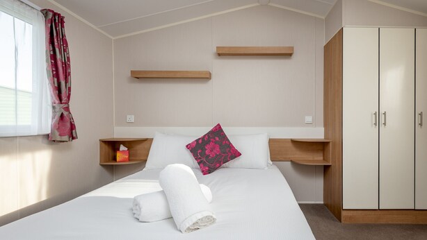 A neatly made bed with white linens and a rolled towel, wooden headboards, a pink floral pillow, and a wardrobe with two doors.