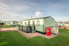 A single-story mobile home with a fenced porch, a small attached shed, and two red gas cylinders.