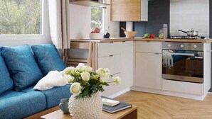 A modern kitchen with white cabinets, a microwave, and a stove. There's a blue sofa with a white cushion and a vase of flowers on a wooden table. - ZEcamping 4 étoiles 500m plage parc aquatique mobil-home 31m2 (Sainte Brevin-les-Pins)