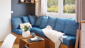 Un salon moderne avec un canapé bleu, un fauteuil blanc et une petite table basse en bois. Des étagères sont placées au-dessus du canapé, où sont disposés des objets décoratifs, et une fenêtre offre une vue sur la verdure à l’extérieur.