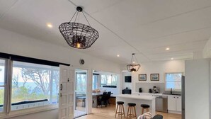 A modern living room with a sofa, coffee table, and chairs, a kitchen area with bar stools, and large windows with a view. - The Gem at Depot Beach (Depot Beach)