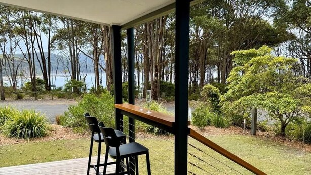 A covered outdoor area with a wooden deck, two black bar stools, and a view of a natural landscape with trees and a body of water. - The Gem at Depot Beach (Depot Beach)