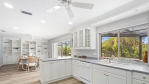A spacious kitchen with white cabinetry, a large window with a view of greenery, and a dining area with a table and chairs.
