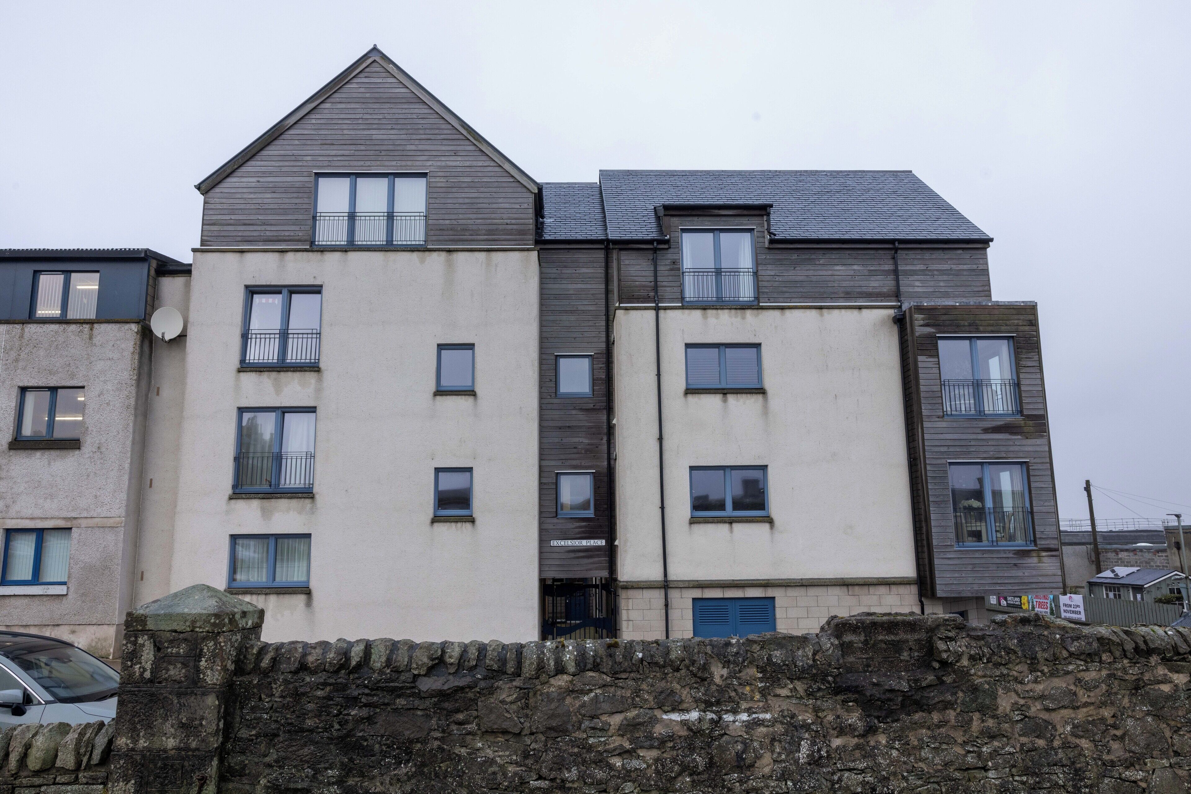 A multi-story residential building with a stone wall in the foreground, featuring a mix of window styles and balconies.