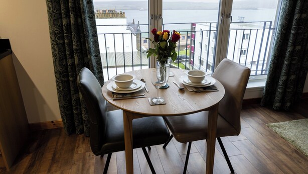 A dining area with a table set for four, a vase with flowers, and a view of a body of water through large windows. - Lerwick Penthouse Apartment (Lerwick)