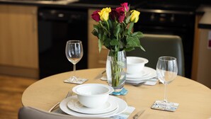 A dining table set with white plates, bowls, cutlery, and two wine glasses. A vase with yellow and red roses is placed in the center. - Lerwick Penthouse Apartment (Lerwick)
