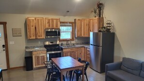 A kitchen with wooden cabinets, a stainless steel refrigerator, a microwave, and a stove. There is a dining table with chairs and a gray sofa in the living area.
