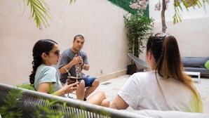 A group of people sitting on outdoor furniture in a patio area with a green wall and a tree with pink flowers. - SUUT Hostal Boutique (Playa del Carmen)