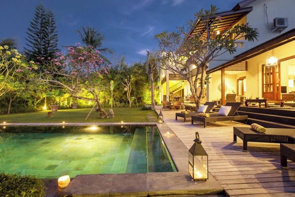 Relax by the serene pool surrounded by lush greenery and flowering plants, with a comfortable lounge area under a shaded pergola.
