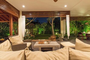 Relax in a serene garden setting surrounded by lush greenery and trees, with a pergola overhead and comfortable wicker seating.