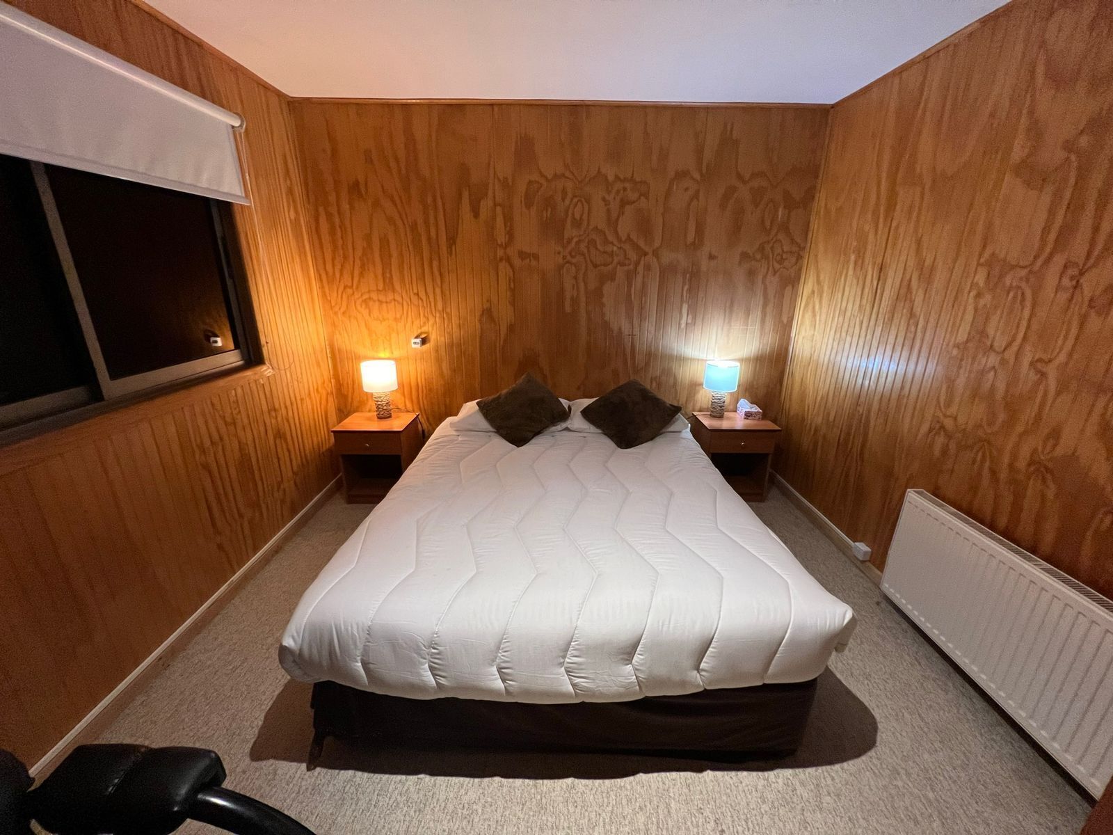 Habitación de hotel con una cama grande, dos mesitas de noche con lámparas, paneles de madera en las paredes y una ventana con persianas.