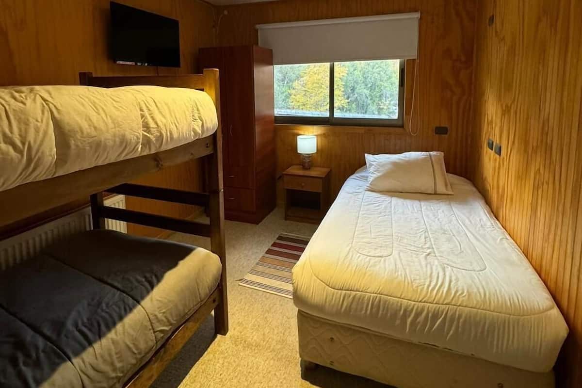 Ein Zimmer mit zwei Etagenbetten, einem Einzelbett, einem an der Wand befestigten Fernseher, einem Fenster mit Blick auf Bäume und einem kleinen Tisch mit einer Lampe.
