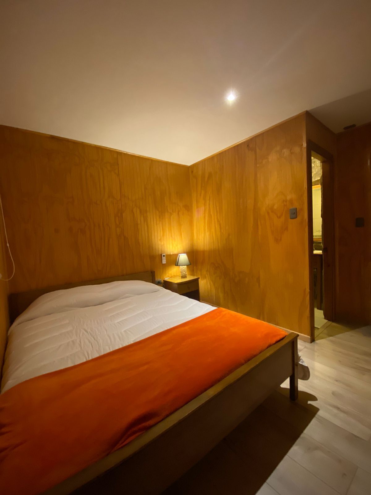 Un dormitorio con cabecera de madera, cama con ropa de cama blanca y naranja, mesita de noche con lámpara y suelo de madera.