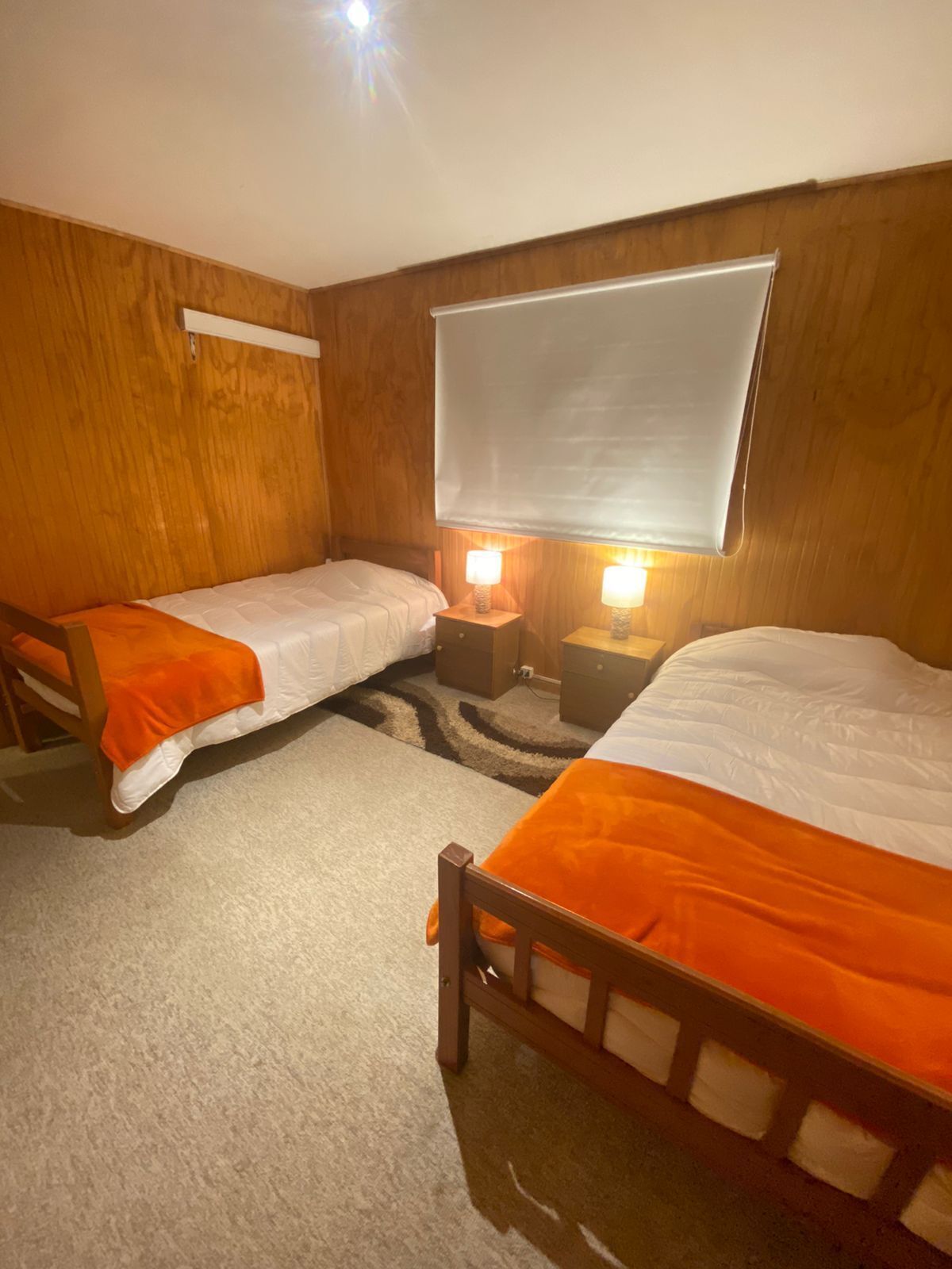 A room with two single beds, wooden walls, a large window with a blind, and bedside lamps.
