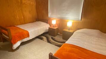 A room with two single beds, wooden walls, a large window with a blind, and bedside lamps.