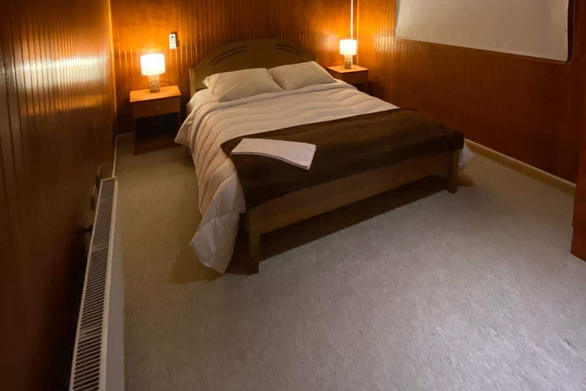 Ein Schlafzimmer mit hölzernem Kopfteil, einem Bett mit weißer Bettwäsche, zwei Nachttischlampen, einer Klimaanlage und einem großen Fenster mit Jalousie.