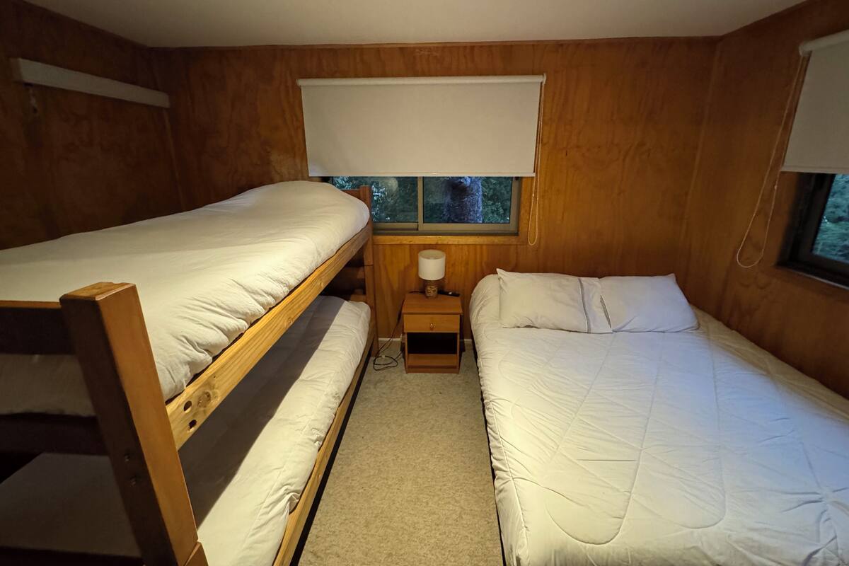 Ein Zimmer mit zwei Etagenbetten, einem Nachttisch mit Lampe und einem Fenster mit Blick auf Bäume.