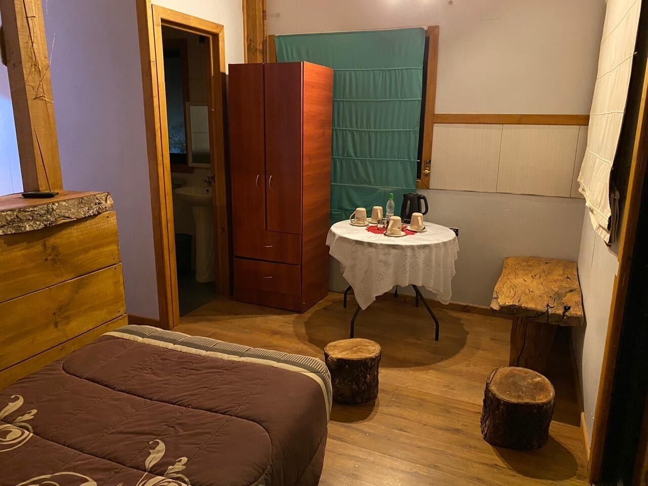 A compact bedroom with a bed, a wooden wardrobe, a small round table with a white tablecloth, and two wooden stools.