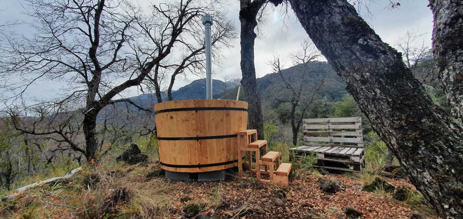 Una struttura in legno rustica con un camino, circondata da alberi e una panchina di legno.