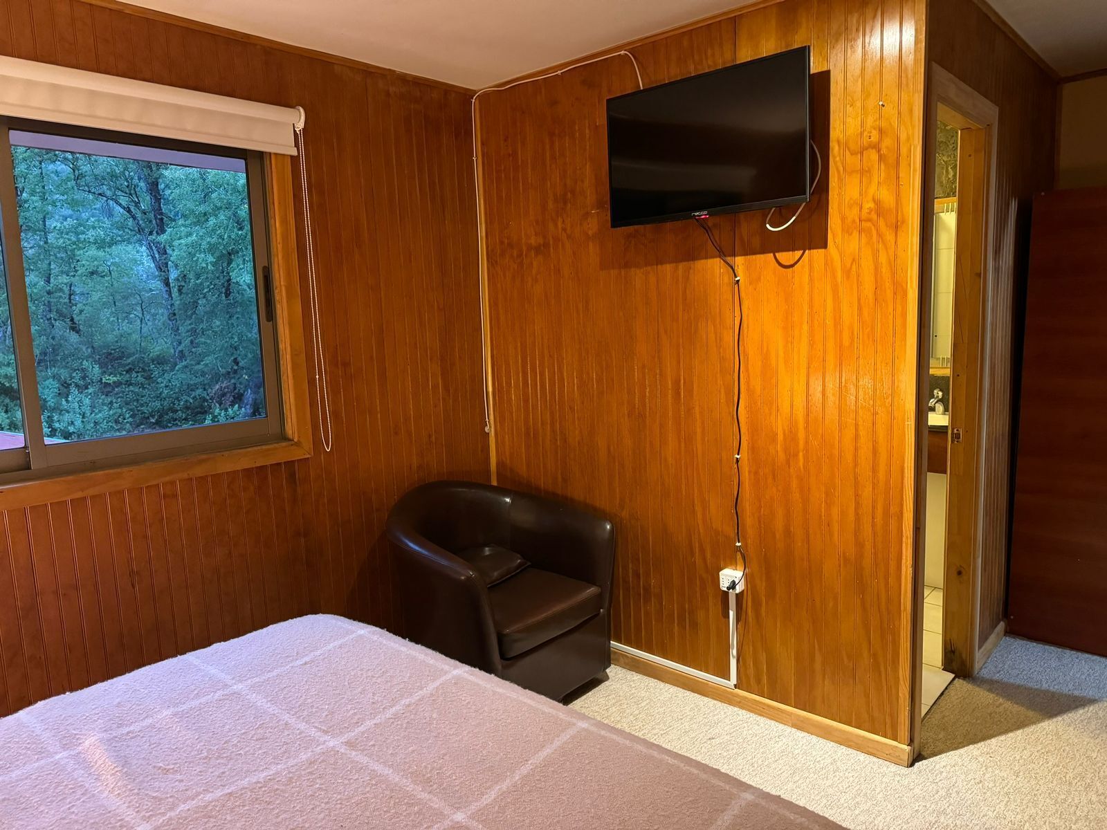 Une pièce aux murs lambrissés, un lit recouvert d’une courtepointe blanche, un fauteuil en cuir noir, un téléviseur fixé au mur et une fenêtre donnant sur la forêt.