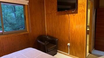 Une pièce aux murs lambrissés, un lit recouvert d’une courtepointe blanche, un fauteuil en cuir noir, un téléviseur fixé au mur et une fenêtre donnant sur la forêt.