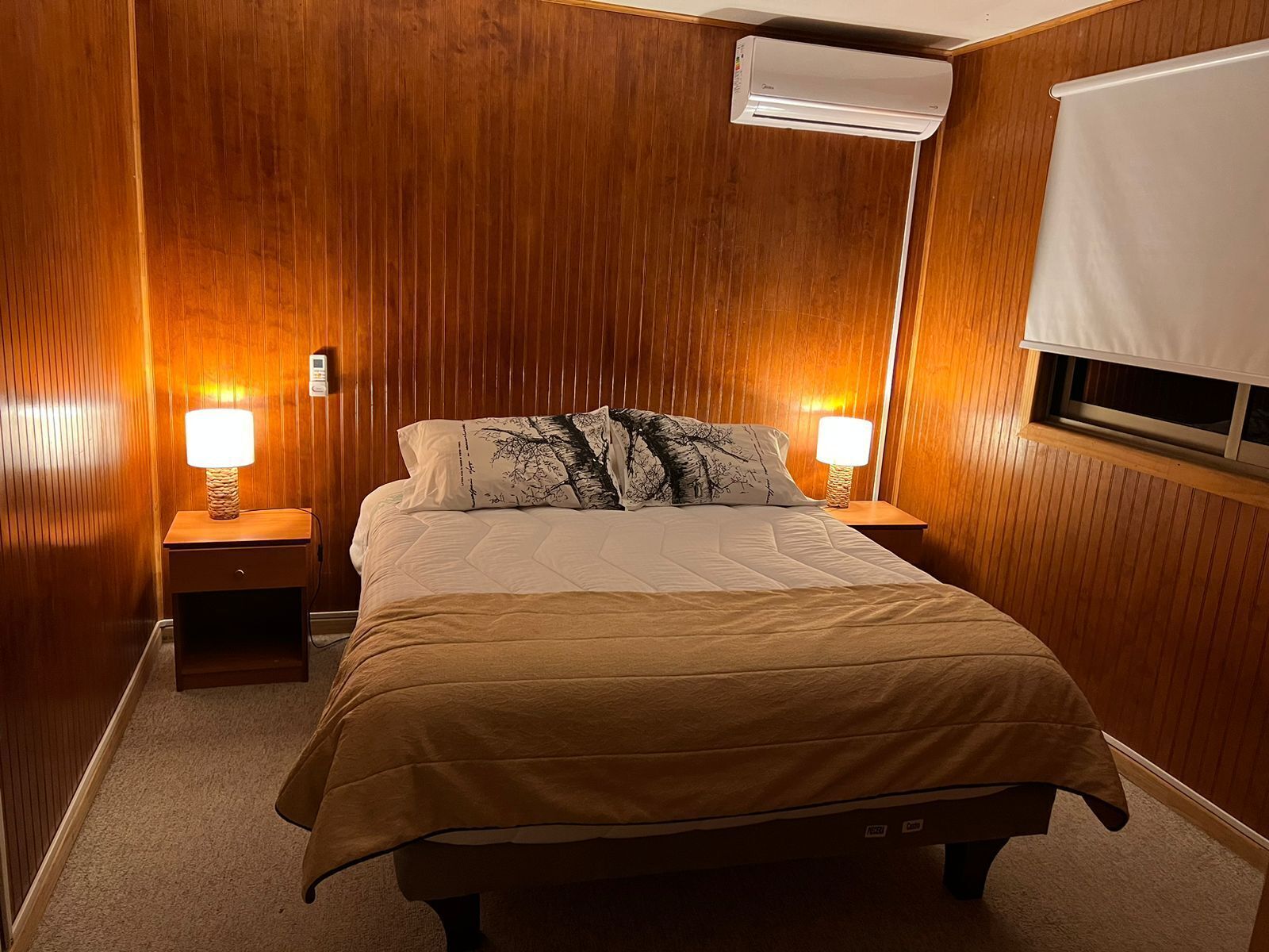 Un dormitorio con pared de paneles de madera, cama con colcha color beige, dos lámparas de noche y aire acondicionado instalado en el techo.
