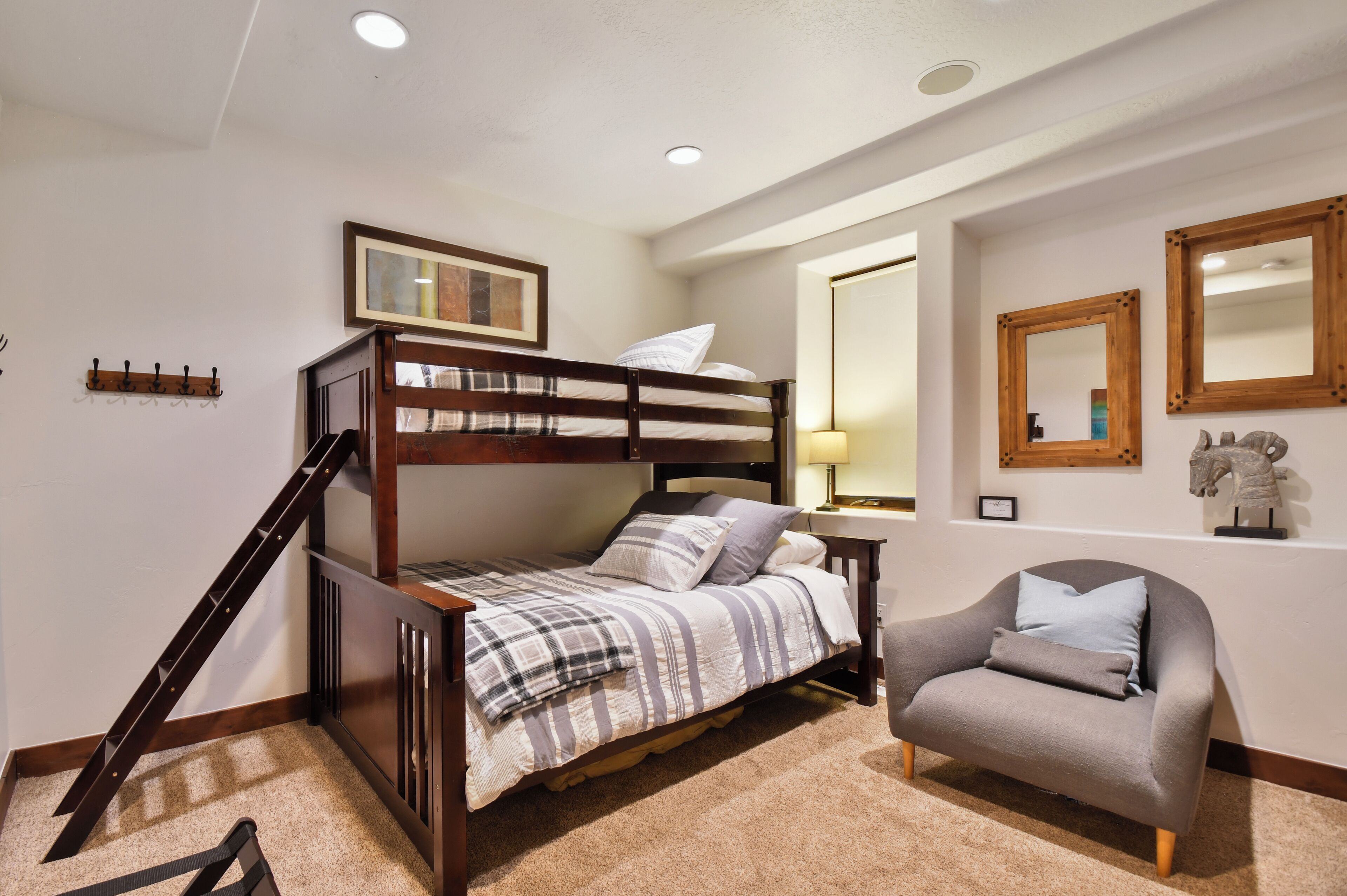 A bunk bed with a wooden frame, a gray armchair, a small table with a lamp, and a mirror on the wall.