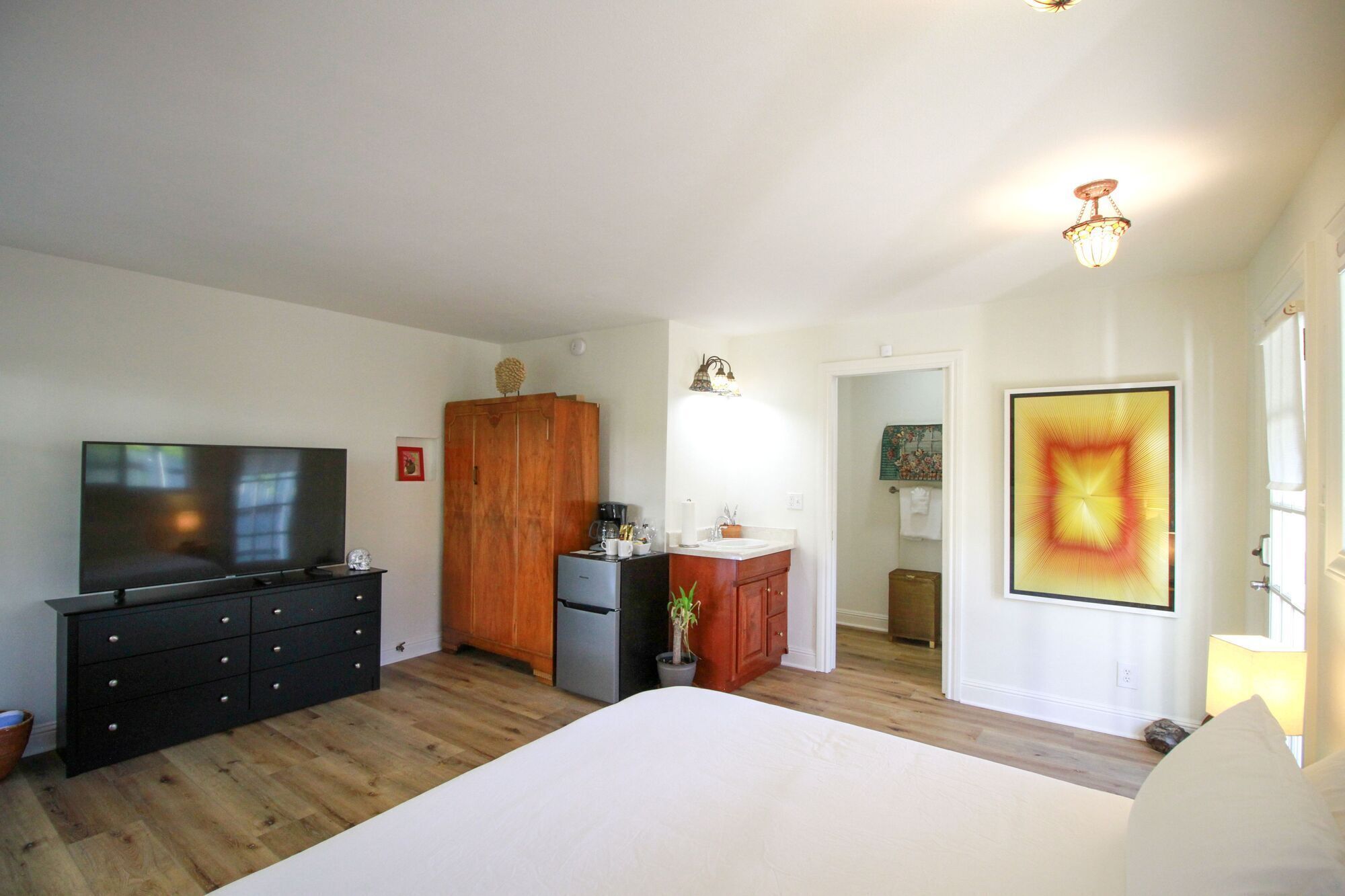 Ein Schlafzimmer mit Bett, Fernseher, Kleiderschrank, Schreibtisch und einem durchgehenden Türrahmen einsehbarer Badezimmer.