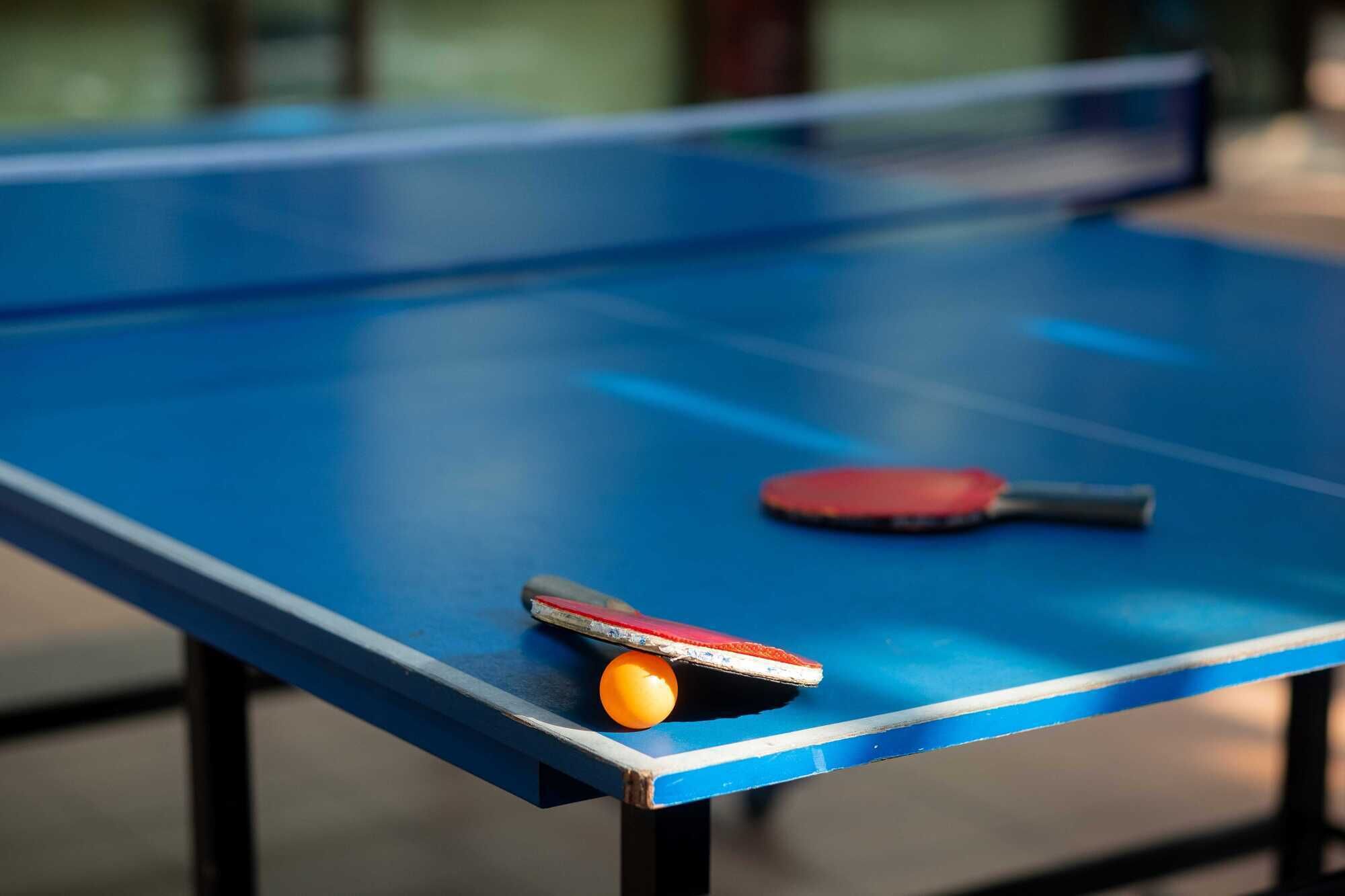 A blue ping pong table with a red and black paddle and a yellow ball.