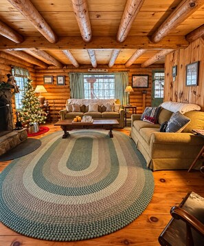 Interior de una cabaña de troncos con techo de madera, alfombra estampada en tonos verde y beige, un sofá, una mesa de centro y un árbol de Navidad.