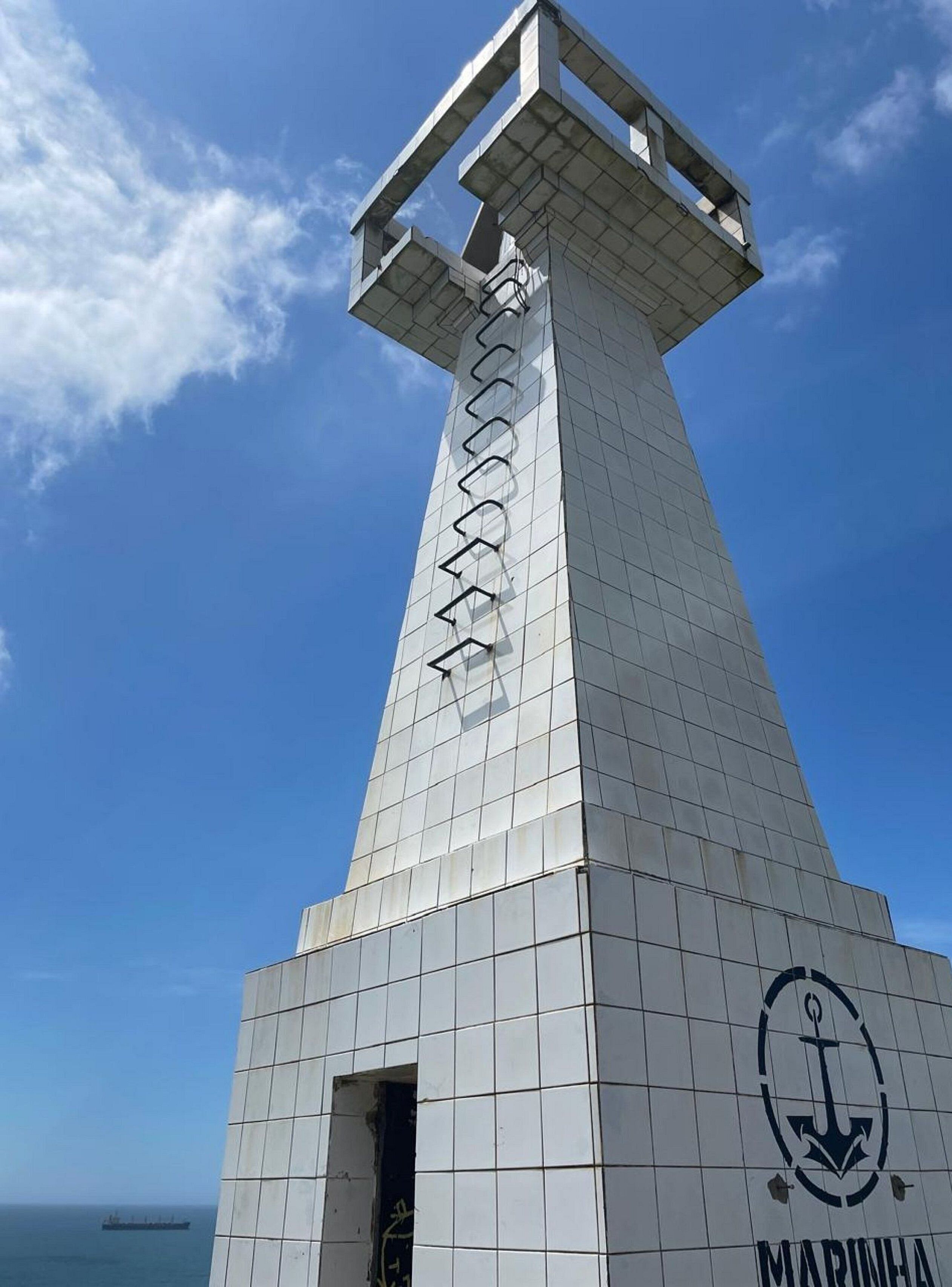 A tall, white, tiled lighthouse with a spiral staircase, an anchor logo, and a view of the sea.
