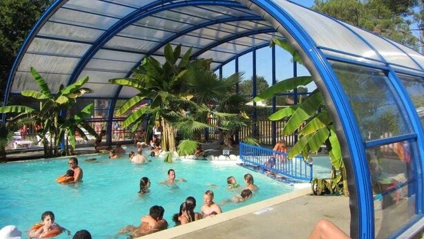 A covered swimming pool with a blue roof, surrounded by palm trees and other greenery. - ZEcamping 4* piscine couverte toboggans locatif 36m2 6 pers (Névez)