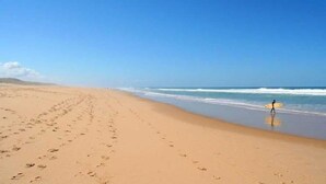 A wide sandy beach with footprints leading to the water, a clear blue sky, and a person with a surfboard.