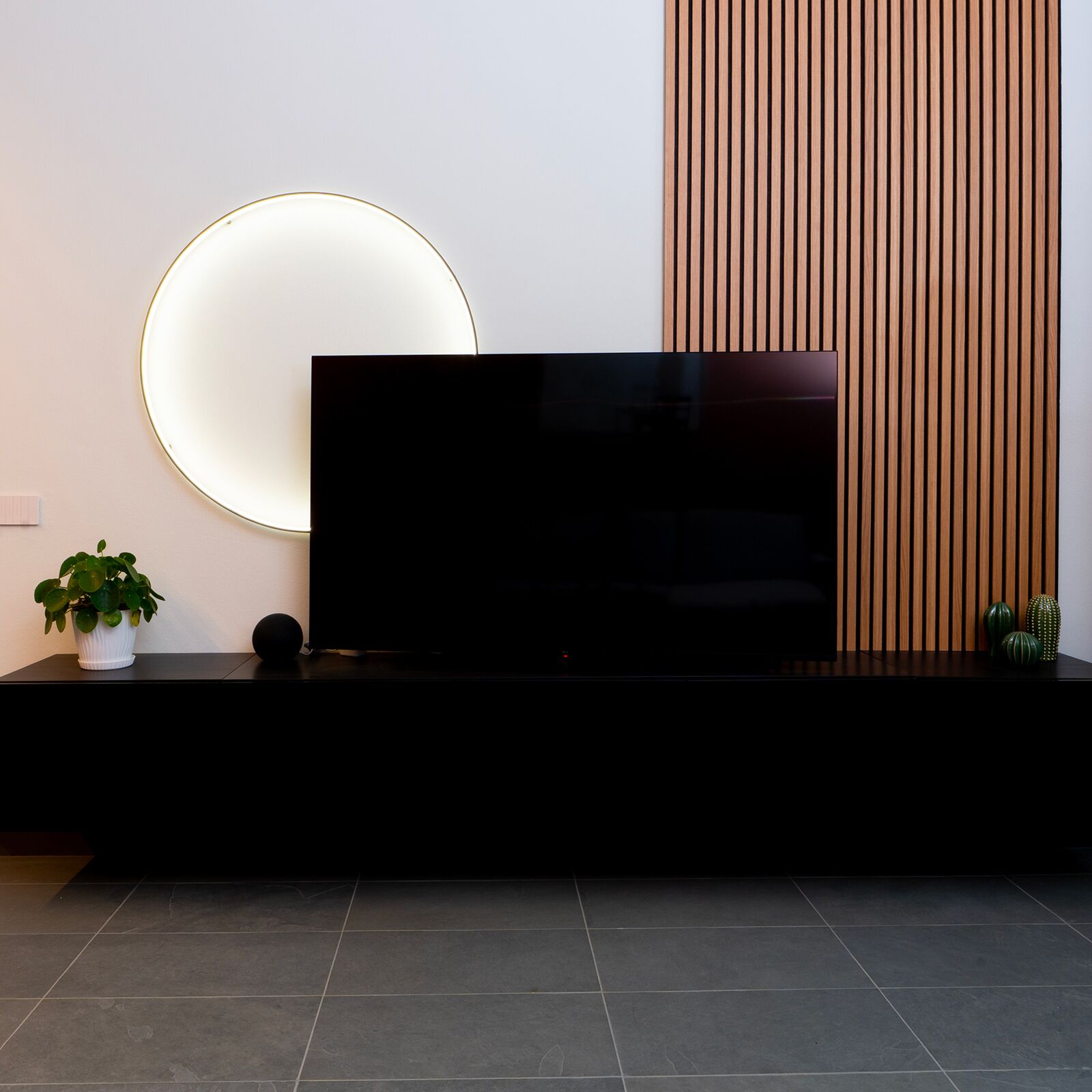 A modern living room with a large flat-screen TV on a black stand, a round wall light, a potted plant, and a wooden panel wall.