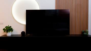 A modern living room with a large flat-screen TV on a black stand, a round wall light, a potted plant, and a wooden panel wall.