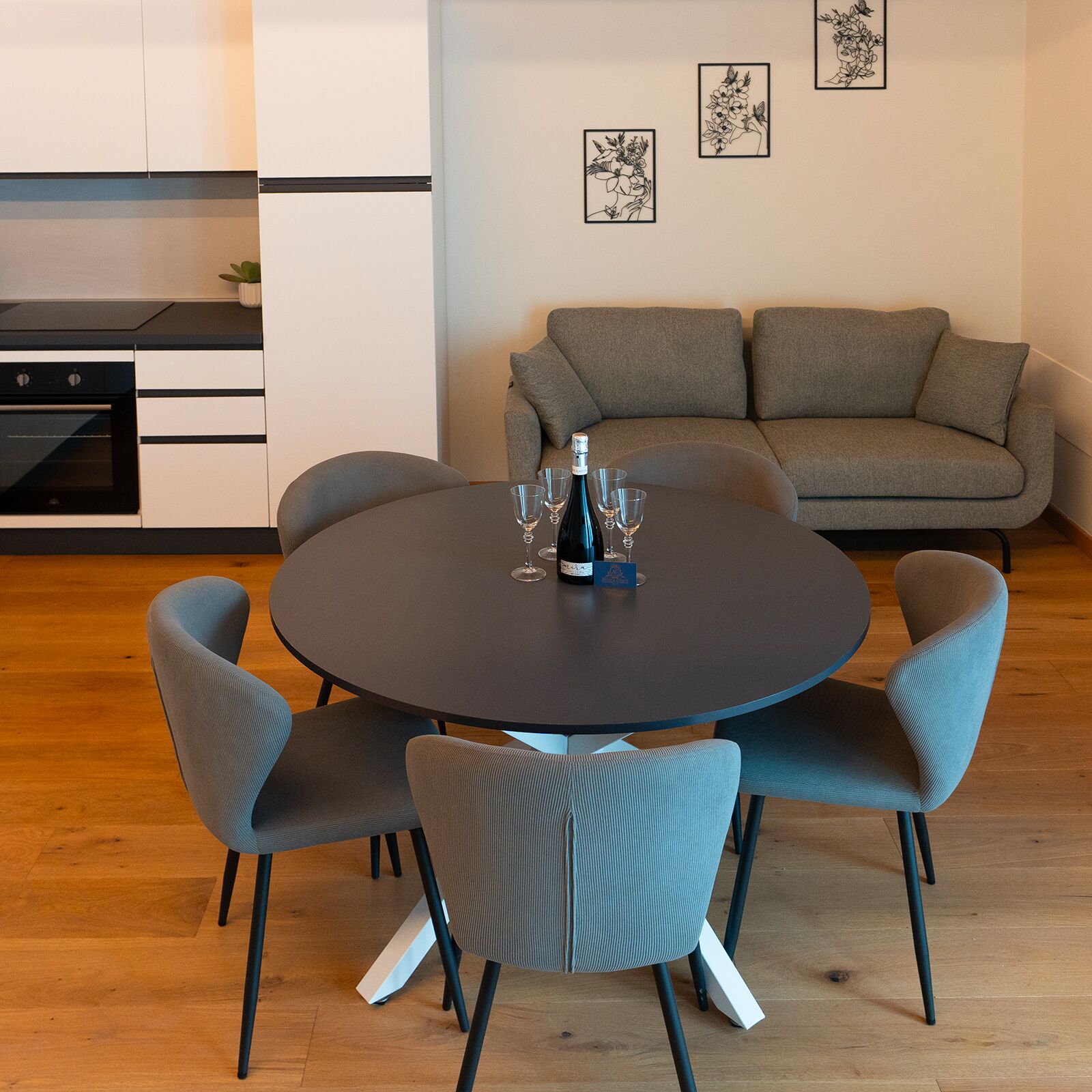 A modern dining area with a round table, grey upholstered chairs, and a sofa in the background.