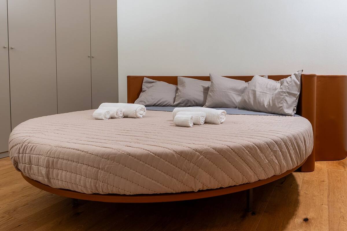 A neatly made bed with a beige quilt and rolled towels, a wooden headboard, and a wooden floor.