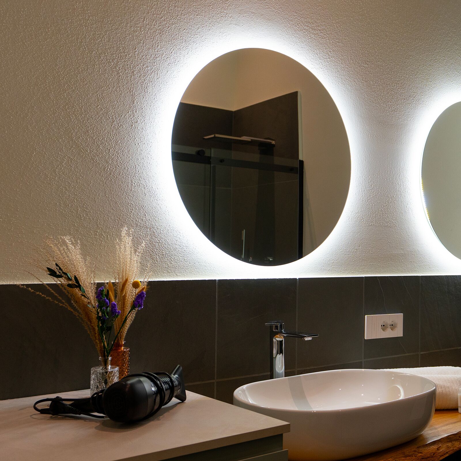 Ein modernes Badezimmer mit einem großen, ovalen Spiegel, einem weißen Waschbecken und einer Duschkabine aus Glas.