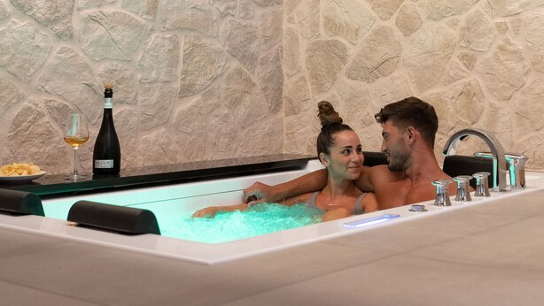 A stone wall, a flat-screen TV, and a bathtub with a bottle and glass. - Antica Corte Lugo (Lugo)