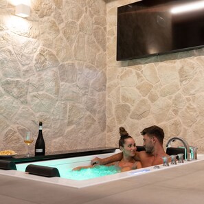 A stone wall, a flat-screen TV, and a bathtub with a bottle and glass. - Antica Corte Lugo (Lugo)