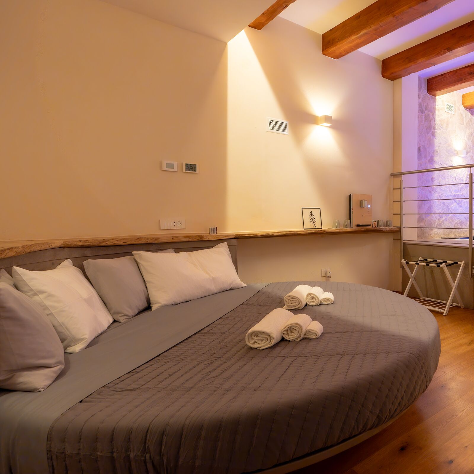A bedroom with a large bed, a wooden ceiling, a stone wall, and a small shelf with a picture frame and a bottle.