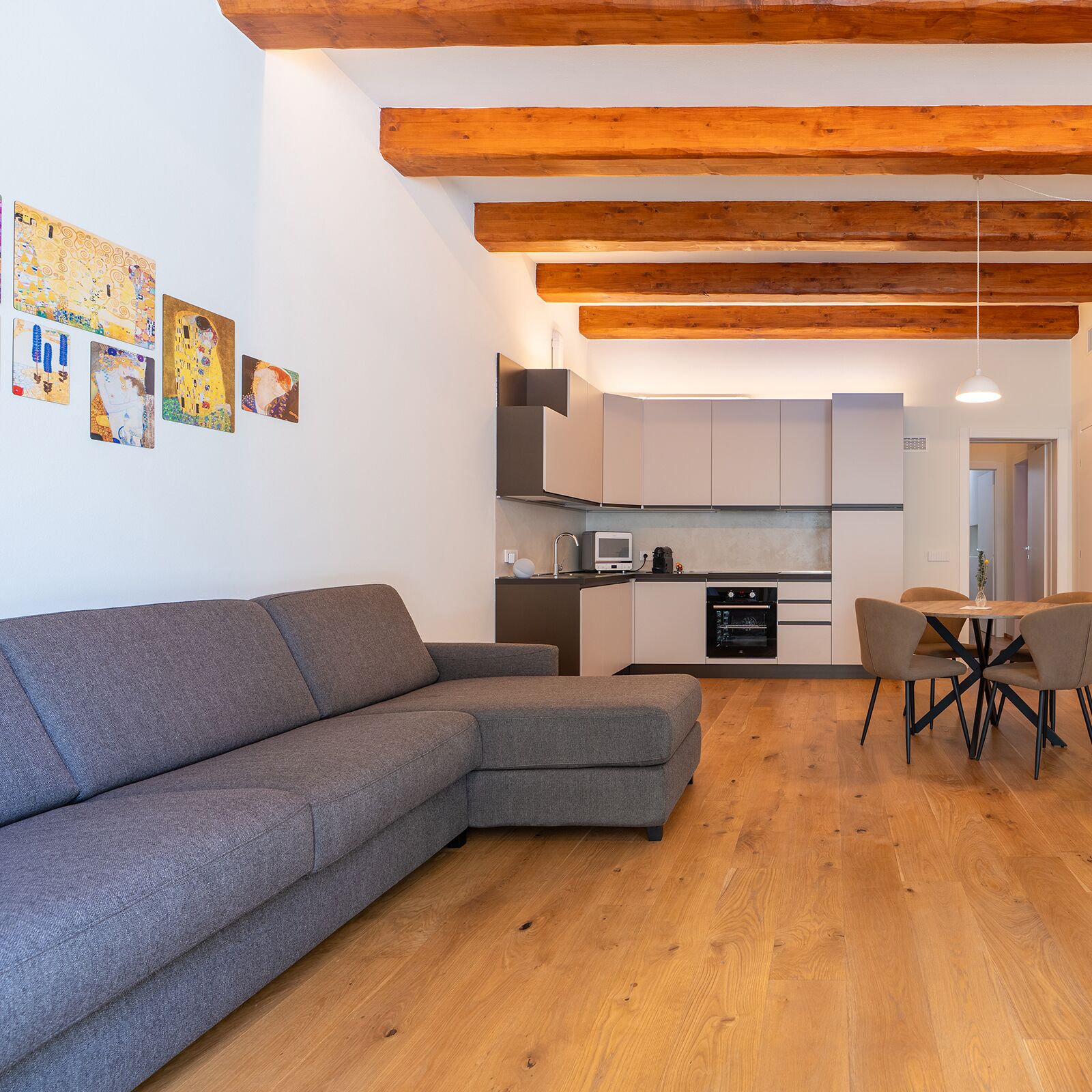 A modern living room with a grey sectional sofa, a dining area with a wooden table and chairs, and a kitchen with white cabinets and a built-in oven.