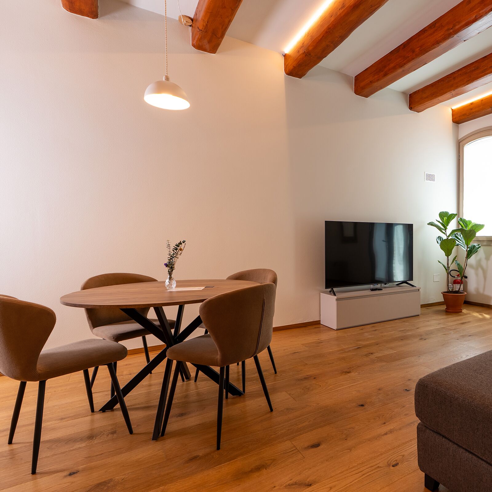Un coin repas avec une table ronde, quatre chaises, un vase de fleurs, un téléviseur à écran plat et une plante en pot.